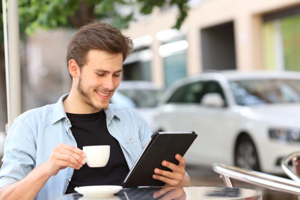 A young man with short brown hair and a beard, wearing a light blue shirt, sits at an outdoor café. He is smiling while holding a white coffee cup in one hand and looking at a digital tablet in his other hand. Cars and a tree are in the background.