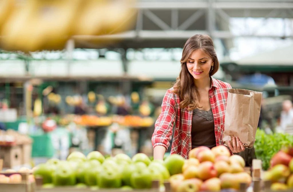 A woman wearing a red plaid shirt holds a paper bag while selecting apples at an outdoor market. She is smiling and focused on choosing the fruit. The market display includes various produce, and there is a lively, bright atmosphere in the background.