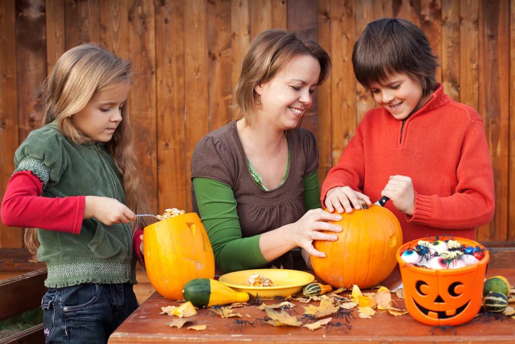 A woman and two children are sitting at a wooden table outdoors, carving pumpkins for Halloween. The table is decorated with leaves, mini gourds, and a small pumpkin-shaped bucket filled with candy.