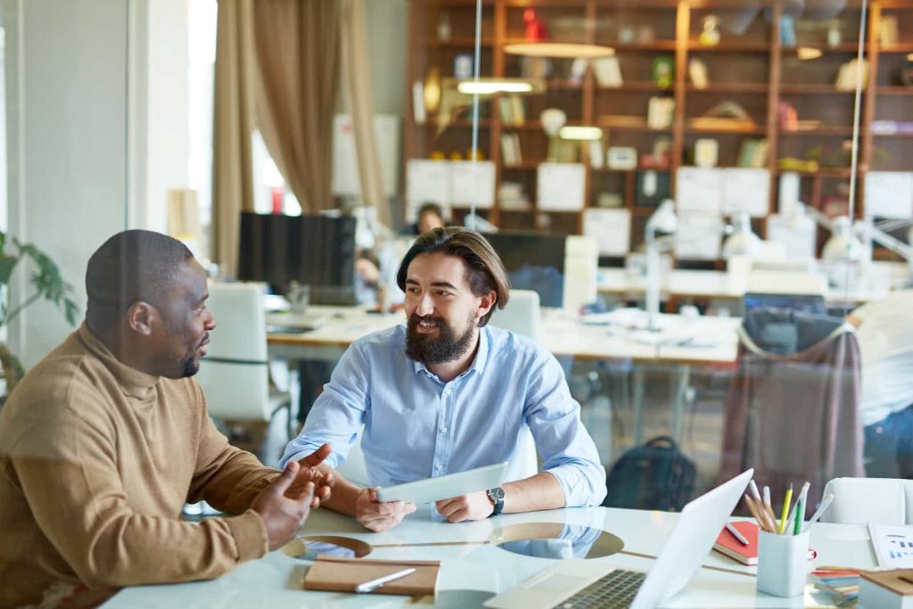 Two men are sitting at a desk in an open-plan office. One is wearing a brown sweater, and the other is in a blue shirt. They are engaged in a conversation, with laptops and papers spread out on the desk. The background shows other desks, computers, and bookshelves.