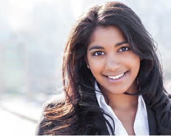 A young woman with long, wavy dark hair smiles warmly at the camera. She is wearing a white collared shirt and a black blazer. The background is softly blurred, suggesting an urban setting.