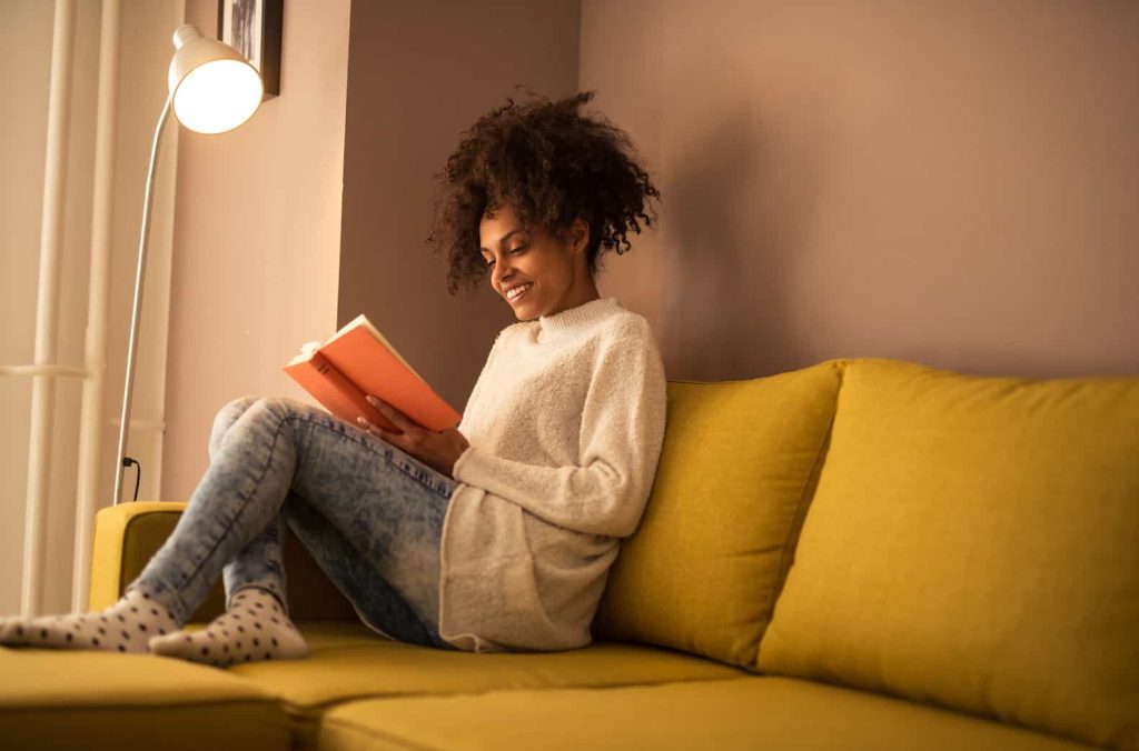A person with curly hair sits on a yellow couch reading a book. They are wearing a cozy white sweater, blue jeans, and white polka dot socks. A standing lamp beside them provides a warm light, creating a cozy atmosphere in the room.