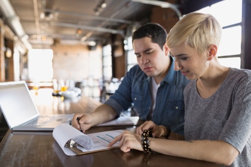 Two people are sitting at a table in a modern workspace. They are collaborating, with the man pointing at a document and the woman looking at it, both focused on the task. A laptop is open on the table. The background features large windows and industrial decor.