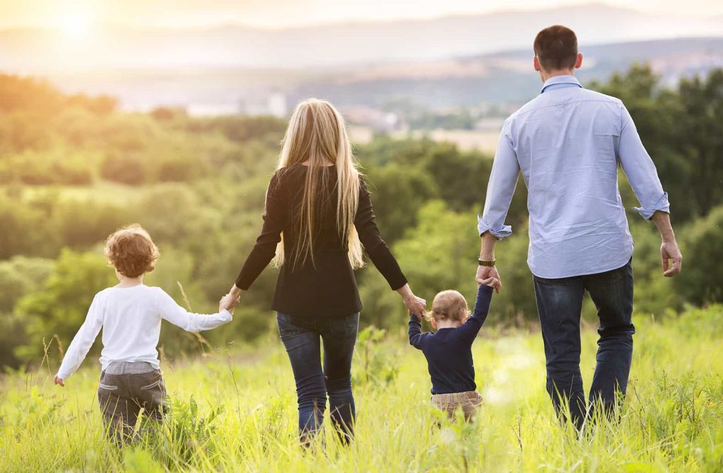 A family of four, including two adults and two children, walks hand-in-hand through a grassy field with picturesque hills and a setting sun in the background. The image is taken from behind, showing the family enjoying a peaceful outdoor moment together.