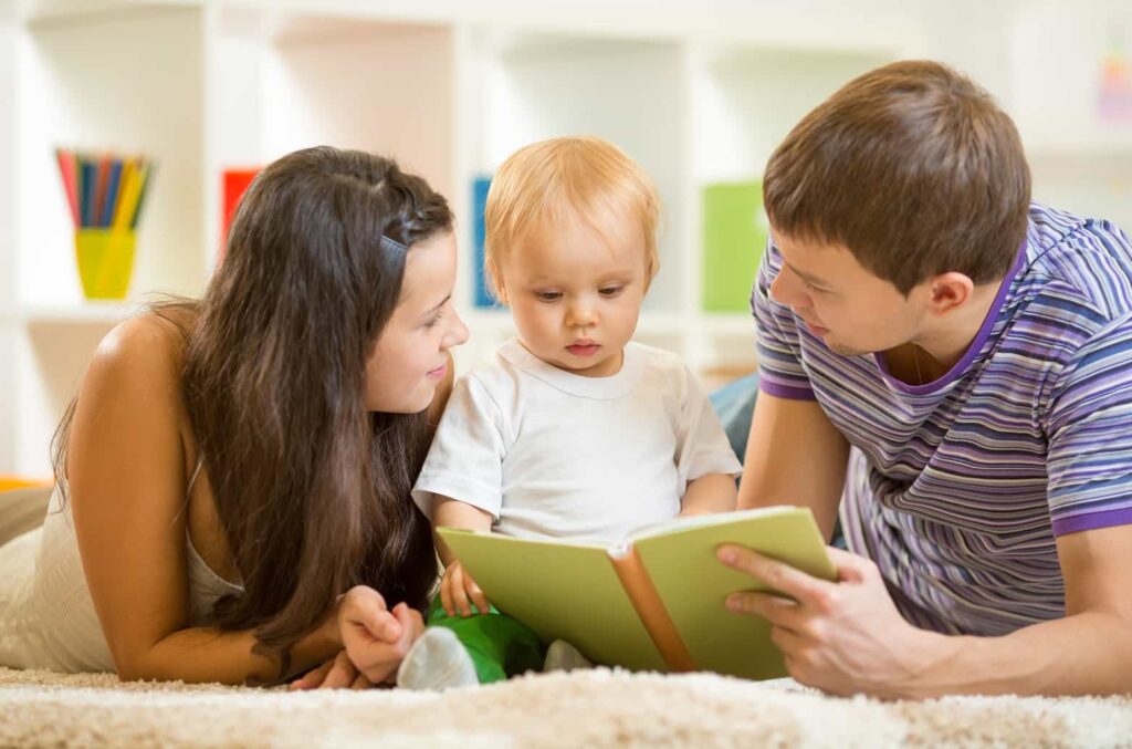 A young child sits between an adult man and woman, all looking at a book the child is holding. They are lying on a soft, carpeted floor with shelves containing colorful books and toys in the background.