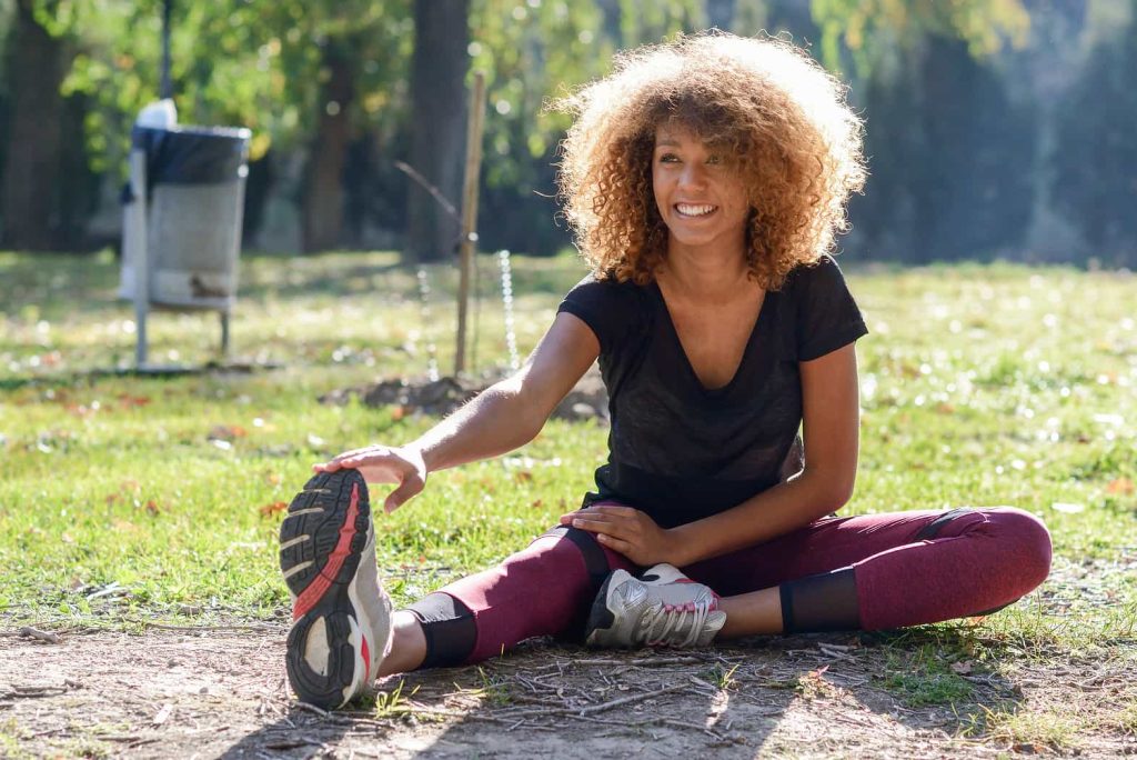 A woman with curly hair sits on the ground in a park, stretching one leg out while touching her sneaker. She is wearing a black t-shirt and red leggings, smiling and looking away from the camera. The park has green grass, trees, and a trash can in the background.