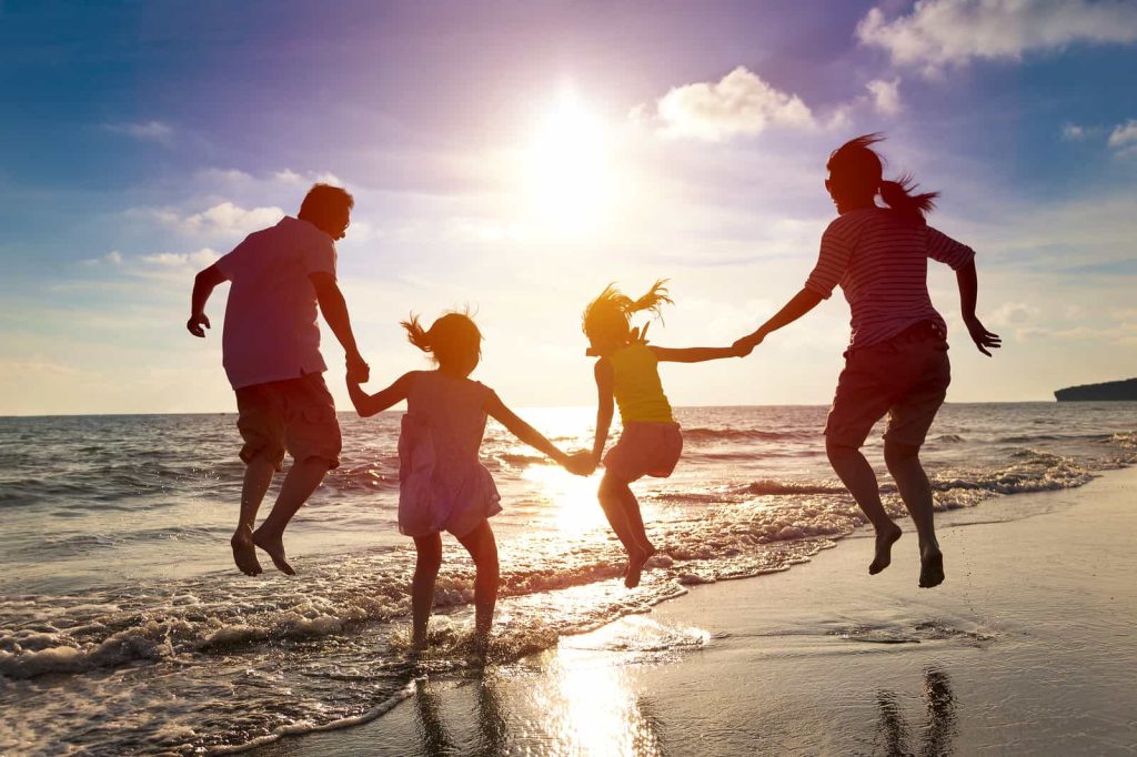A family of four, silhouetted by the setting sun, holding hands and jumping along the shoreline of a beach. The two adults are on either side, with the two children in the middle. The sky is vibrant with hues of orange and purple, reflecting on the water.