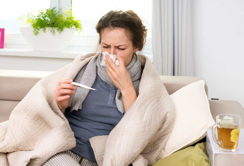 A woman wrapped in a blanket sits on a couch, holding a thermometer in one hand and using a tissue to blow her nose with the other. She appears to be sick. A cup of tea and a box of tissues are on a table next to her.