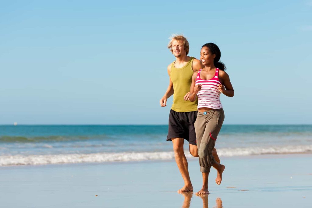 A man and a woman are jogging along the shoreline of a beach on a sunny day. The man is wearing a yellow tank top and black shorts, while the woman is in a striped tank top and cropped khaki pants. Both are barefoot and smiling as they run near the water.