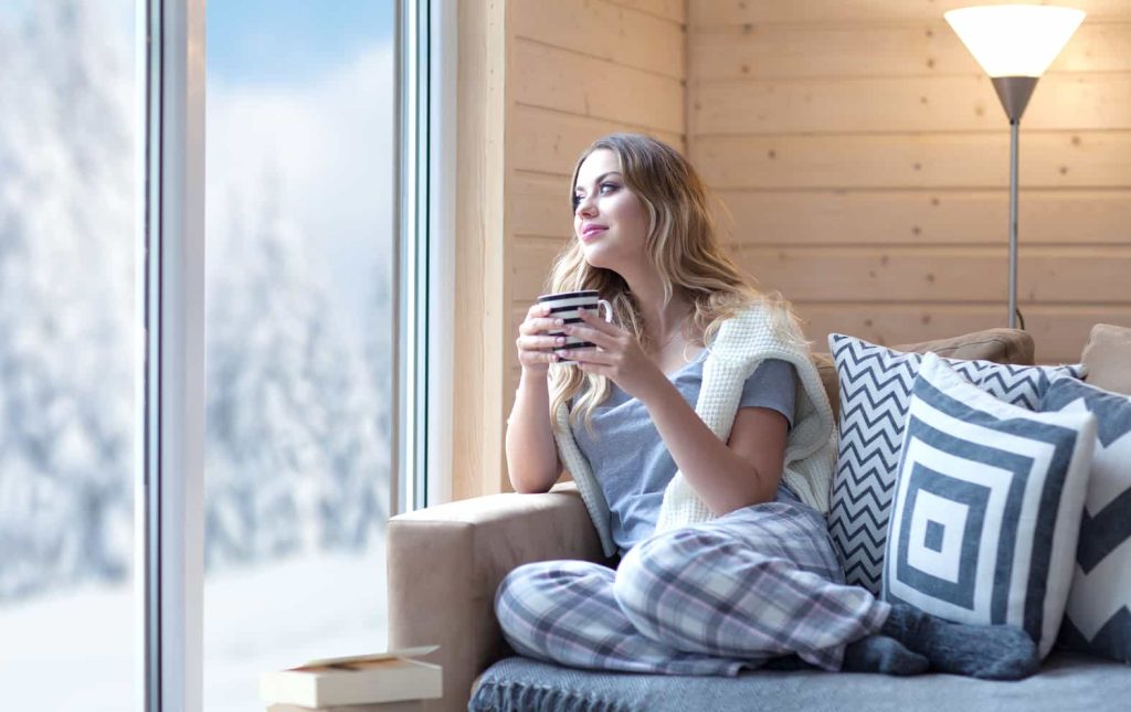 A woman in cozy pajamas and a sweater sits on a couch near a large window, holding a mug. She looks out at a snowy landscape with evergreen trees. The room has wooden walls and a floor lamp, creating a warm and inviting atmosphere. A book lies beside her.