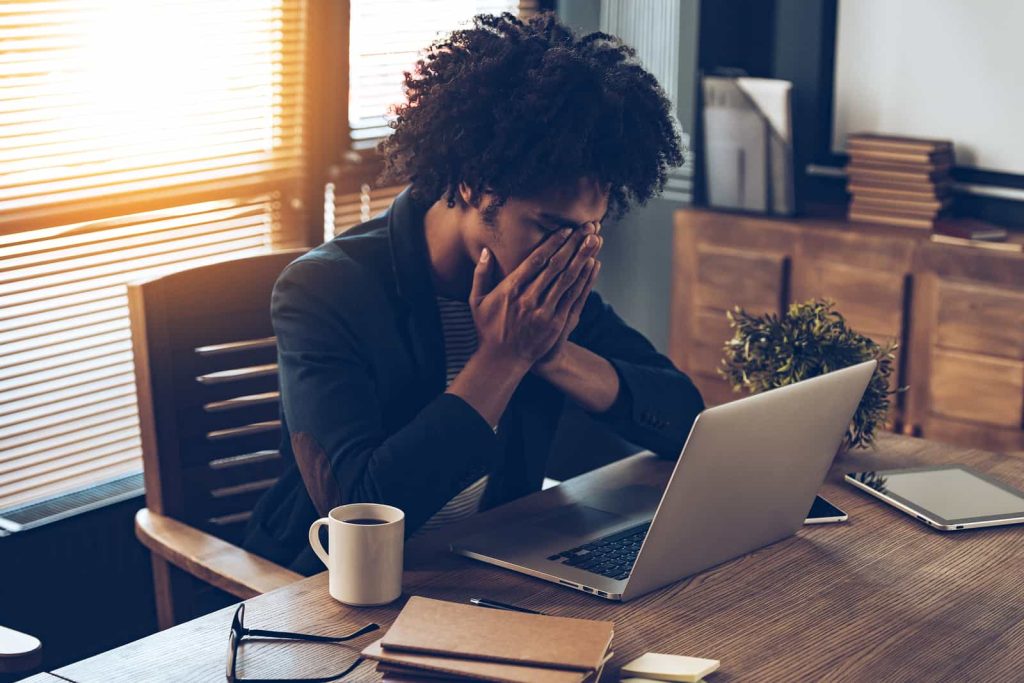 A person sitting at a desk, holding their face with both hands in a gesture of frustration or stress. A laptop, coffee mug, notebook, glasses, and plant are on the desk.