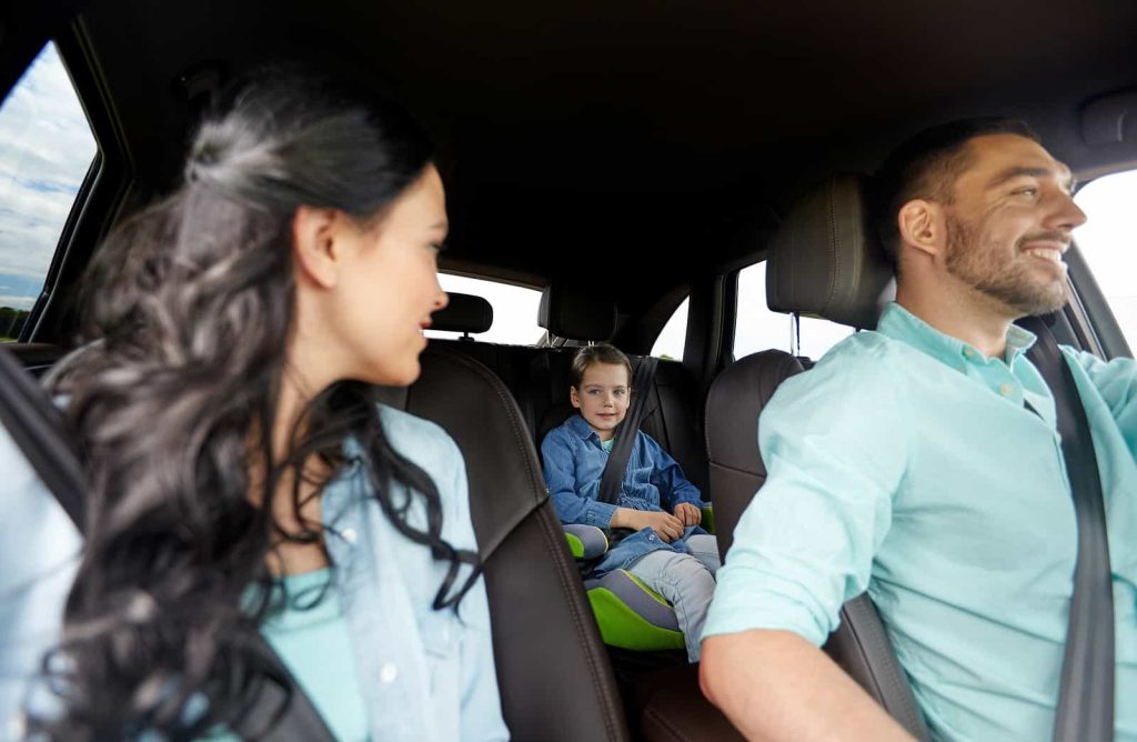 A family in a car: a woman with long dark hair smiles at a child in the back seat, who is wearing a seatbelt and smiling back. A man with short dark hair, also smiling, is driving.