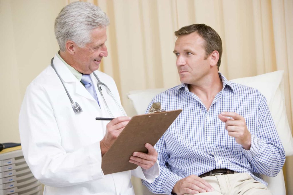 A doctor in a white coat holding a clipboard and pen is speaking with a man in casual attire sitting on an examination table. The doctor is smiling and attentive while the patient gestures with his right hand, engaged in the conversation.