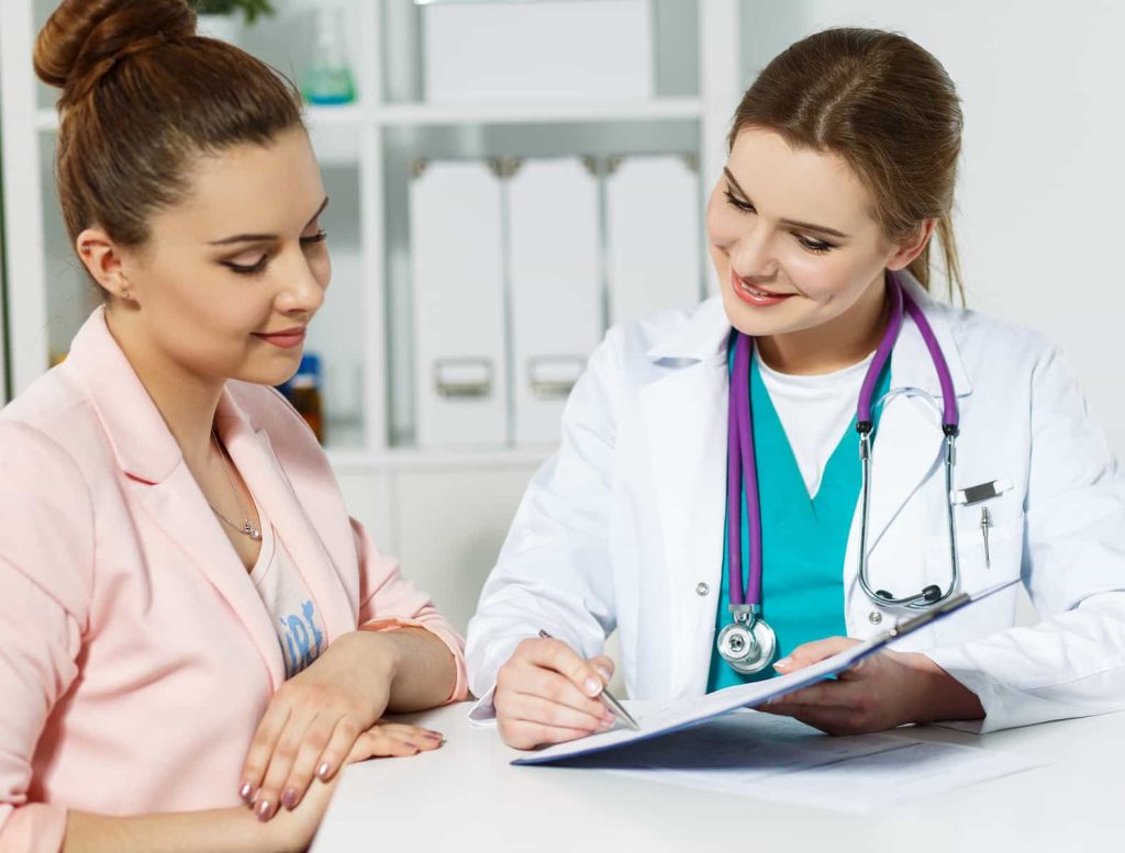 A female doctor in a white coat with a stethoscope around her neck is sitting at a desk and talking to a female patient. The doctor is writing on a clipboard and smiling, while the patient, dressed in a light pink blazer, looks on attentively.