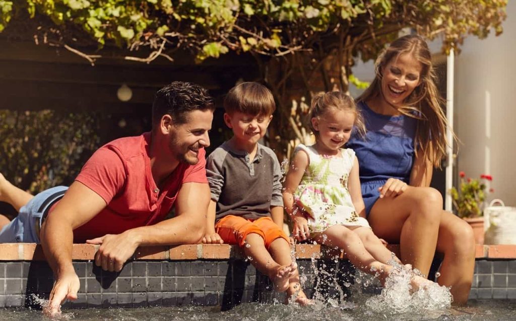 A happy family of four, with parents and two young children, sit on the edge of a pool, splashing their feet in the water. The parents smile while the children, a boy and girl, look excited and playful. Trees and plants are visible in the background.