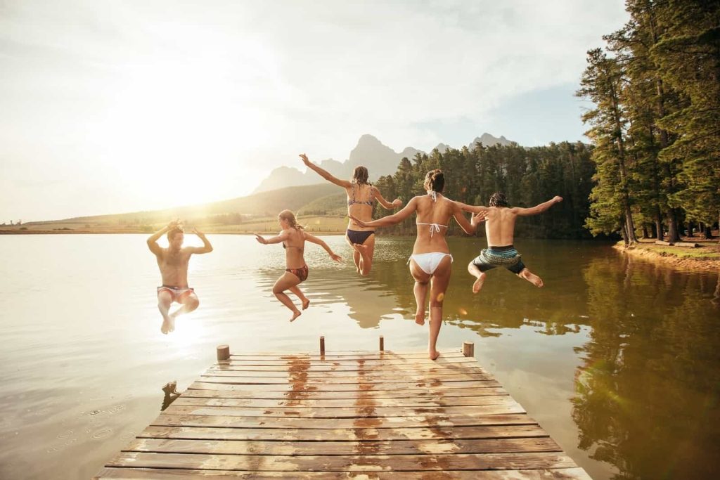 Five people in swimwear are jumping off a wooden dock into a calm lake. They are holding hands or mid-air in various dynamic poses. The sun is setting in the background, casting a warm glow over the mountains and trees surrounding the lake.