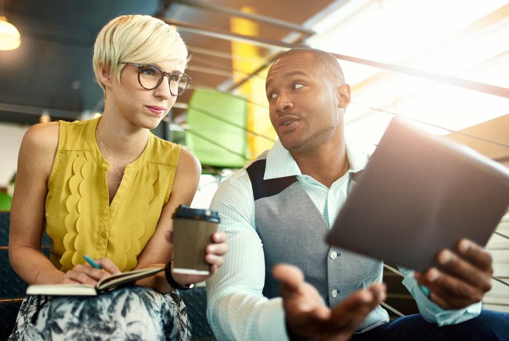 A man and a woman are having a discussion. The man, holding a tablet, gestures with his hand while speaking. The woman, holding a coffee cup and a notebook, looks at him attentively, with a pen in her hand. They appear to be in a modern office setting.