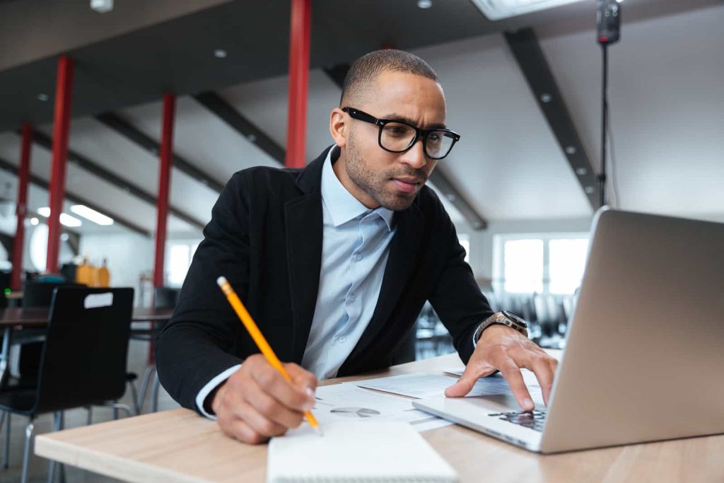 A man wearing glasses is focused on his laptop while holding a pencil and taking notes in a notebook. He is seated at a table in a modern, open workspace with red columns and large windows visible in the background.