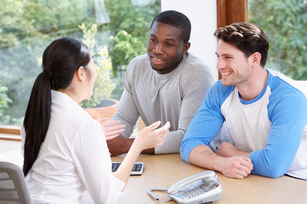 Three people having a discussion at a table. A woman is speaking while two men listen attentively. A phone and papers are on the table, and there is a window with greenery in the background.