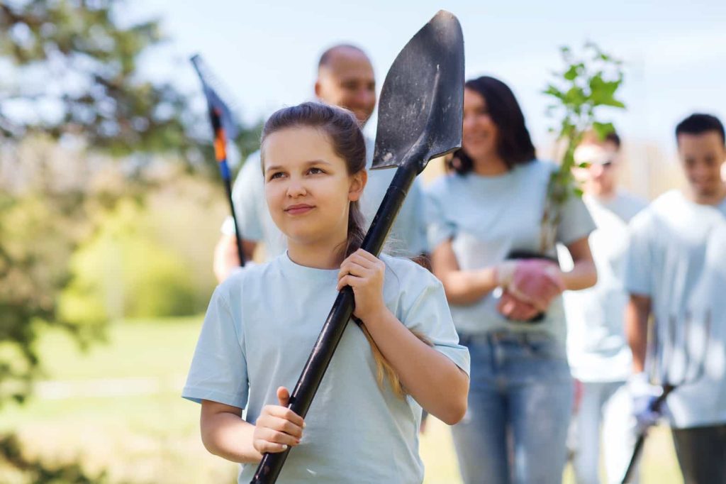 A young girl in a light blue t-shirt holds a shovel while standing outdoors. Behind her, a group of people also in light blue shirts are blurred, holding various planting tools and a small tree. They appear to be participating in a community gardening or tree-planting event.