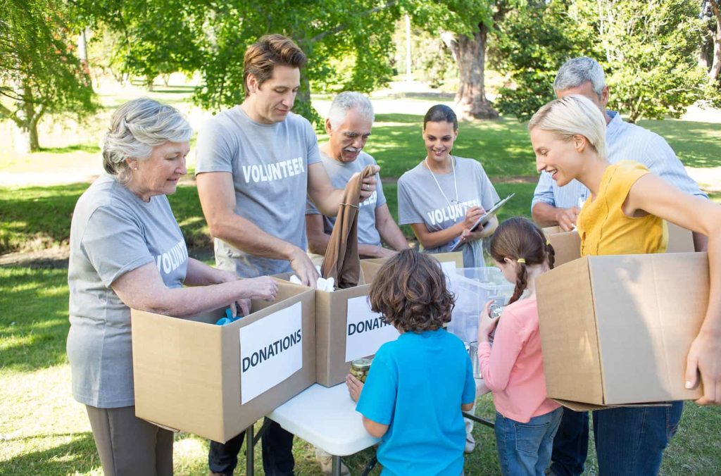 A group of volunteers of varying ages are outdoors, sorting donations into large cardboard boxes labeled "DONATIONS." They are smiling and appear engaged in the activity. .