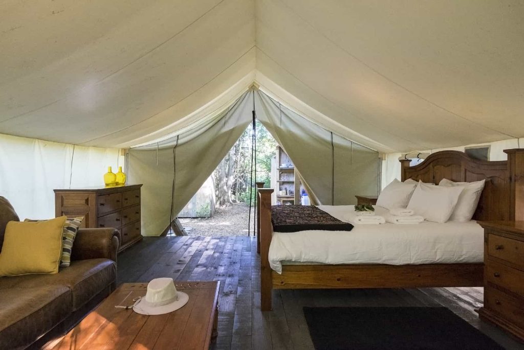 A spacious glamping tent with a cozy interior featuring a large bed with white bedding, wooden nightstands, a brown leather sofa, and a wooden coffee table. The tent is open at the entrance, revealing a view of trees and nature outside.