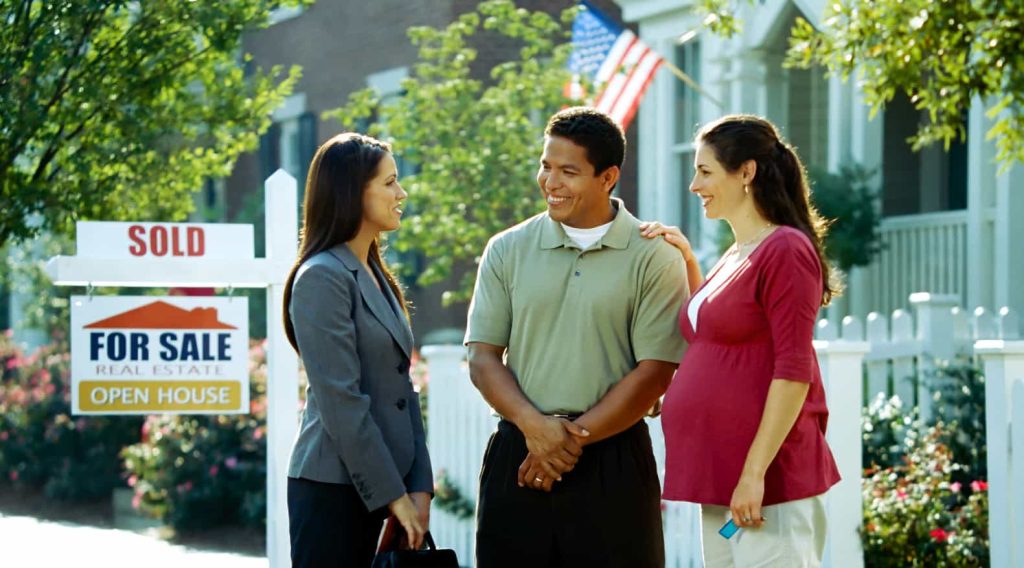 A real estate agent stands with a smiling couple in front of a house with a "Sold" sign. The woman in the couple is pregnant and holds her partner's hand, while the house with an American flag are visible in the background.