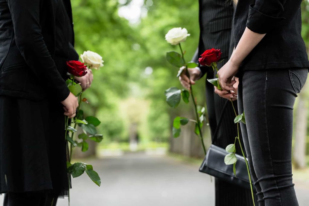 Four people dressed in black, each holding a white or red rose, stand together on a path in a green, leafy setting. The somber mood suggests they are attending a memorial or funeral.