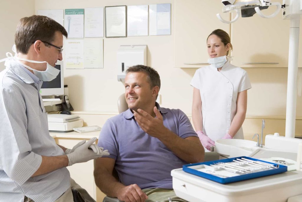 A male dentist wearing glasses, a mask, and gloves is talking to a smiling male patient sitting in a dental chair. A female dental assistant in a white uniform stands nearby. Dental instruments are visible in the foreground.