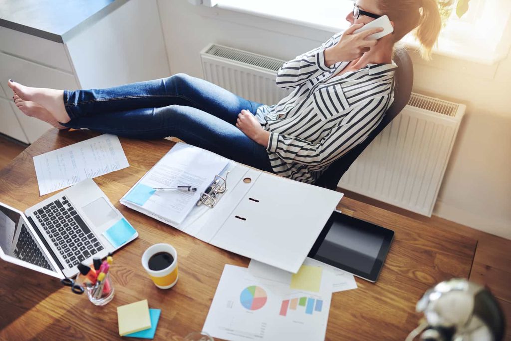A woman is working from home, talking on the phone with her feet up on the desk. The desk is cluttered with a laptop, documents, a clipboard, a cup of coffee, colorful pens, and charts. She appears relaxed and focused on her call.