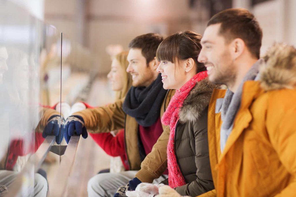 A group of four people, two men and two women, dressed in winter clothing stand side by side, smiling and looking ahead. They are lined up close to a glass barrier, possibly watching an event.