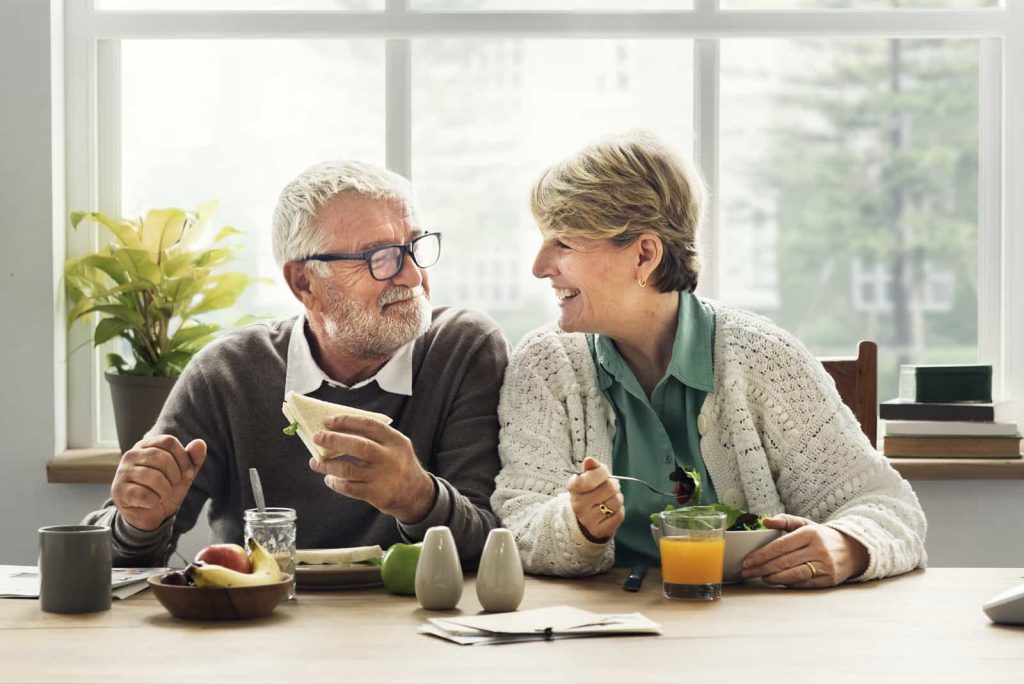 A senior couple is sitting at a kitchen table, smiling at each other while enjoying a meal. The man is holding a sandwich, and the woman is holding a fork. There are plates of food, fruits, and a glass of orange juice on the table.