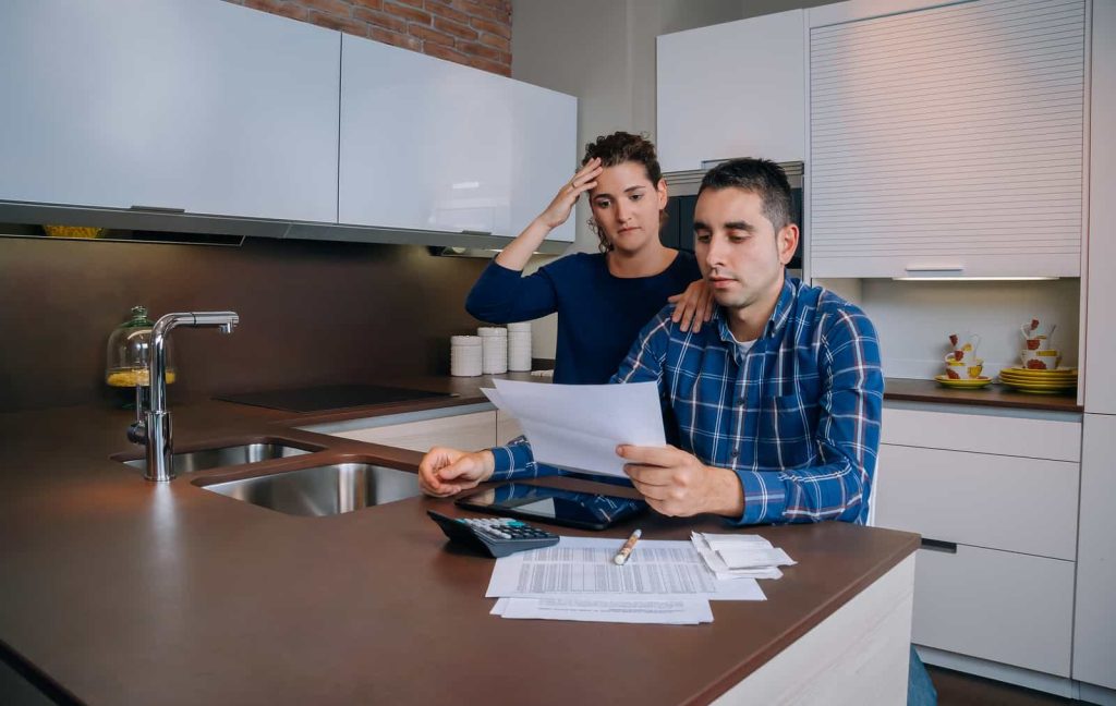A couple in a modern kitchen reviews documents at a countertop. The man in a blue plaid shirt holds a piece of paper, looking concerned, while the woman beside him, wearing a blue sweater, rests her hand on his shoulder with a worried expression.