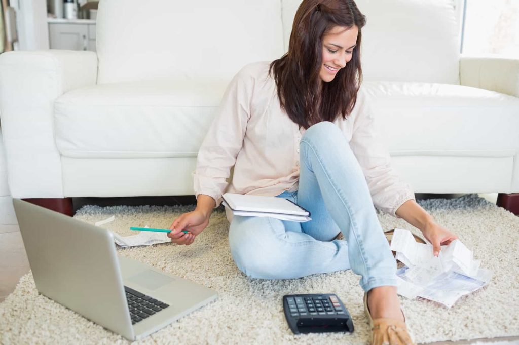 A woman sitting on the floor in front of a white sofa, working with documents. She is holding a piece of paper in one hand and a notebook in the other. A laptop, a calculator, and other papers are spread out around her. She appears focused and smiling.