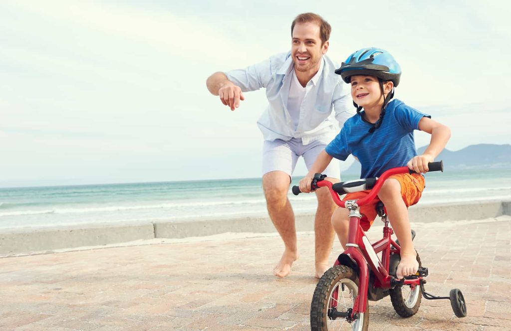 A smiling father encourages a young child, who is riding a red bicycle with training wheels. The child is wearing a blue helmet and blue shirt with orange shorts. They are near the ocean on a paved walkway, with waves and mountains visible in the background.