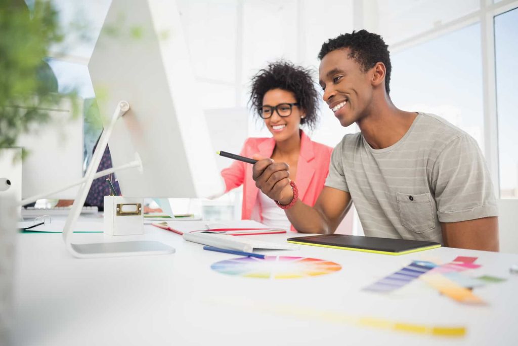 Two people sit at a bright, modern desk, looking at a large computer monitor. One holds a pen and tablet, while the other smiles, wearing glasses. Color swatches and design tools are on the desk, suggesting a creative project or graphic design work.