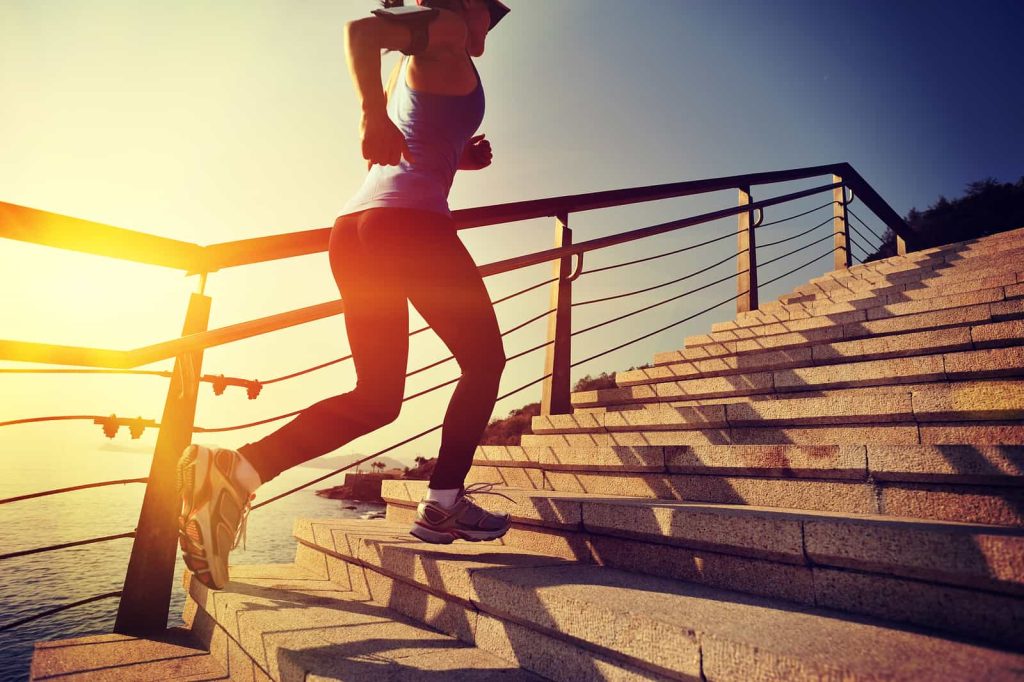 A woman in athletic wear is running up a set of outdoor stairs beside a body of water at sunset. She is wearing a tank top, leggings, and a cap, with her shadow visible on the stairs. The scene is bathed in warm, golden light.
