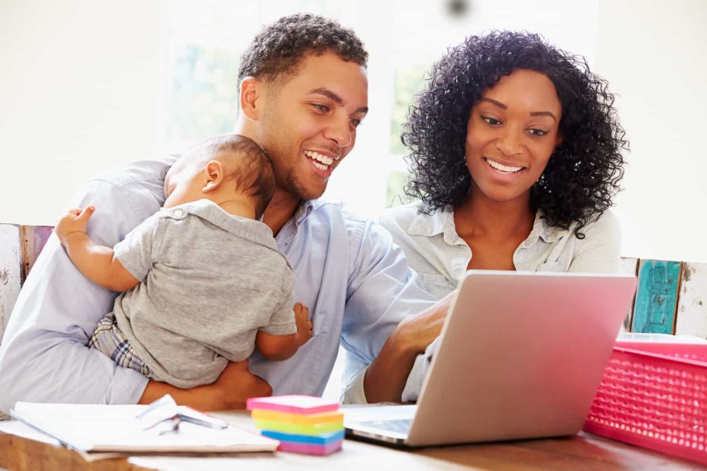 A smiling couple sits at a table looking at a laptop. The man is holding a baby who is resting on his shoulder. On the table are sticky notes, documents, and a pink storage box. Bright natural light streams in from a nearby window.