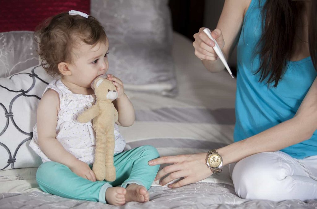 A small child with curly hair sits on a bed holding a stuffed bear. They are looking at an adult, only partially visible, who is holding a digital thermometer.