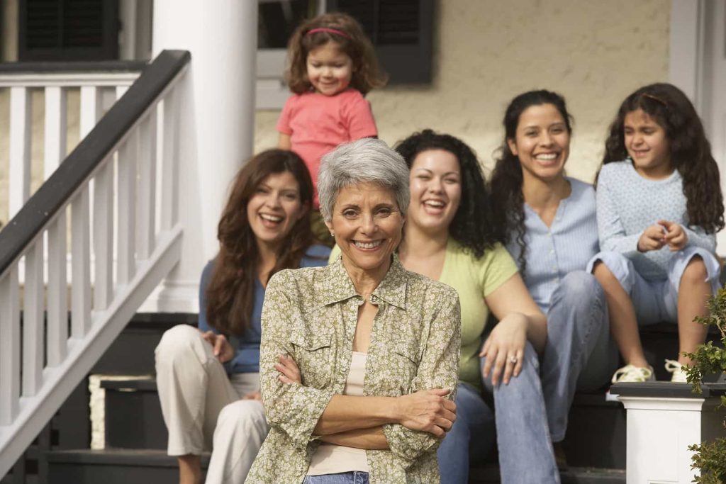 Five women of different ages are smiling in front of a house. An elderly woman with silver hair is standing in front, while the other women sit on the steps behind her. A young girl with curly hair and a red dress stands at the top of the stairs. They all look happy.