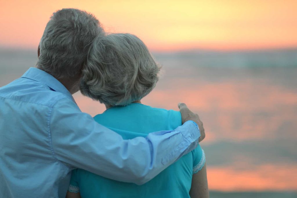 An elderly couple, viewed from behind, gazes at a serene sunset over the ocean. The man, in a light blue shirt, has his arm around the woman, who is wearing a turquoise top, capturing a tender and peaceful moment as the sky glows in warm hues.