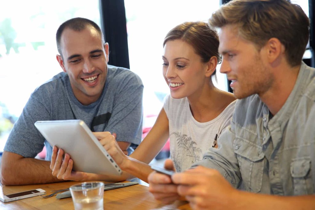 Three people sit at a wooden table looking at a tablet. A woman holds and points at the tablet while two men, one on each side, observe with interest and smile. The setting appears to be a casual indoor location with large windows in the background.