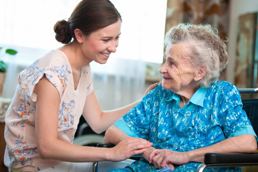 A young woman with brown hair, wearing a light floral blouse, leans down and smiles at an elderly woman sitting in a wheelchair. The elderly woman, with grey hair and wearing a blue patterned dress, holds the young woman's hand while smiling back.
