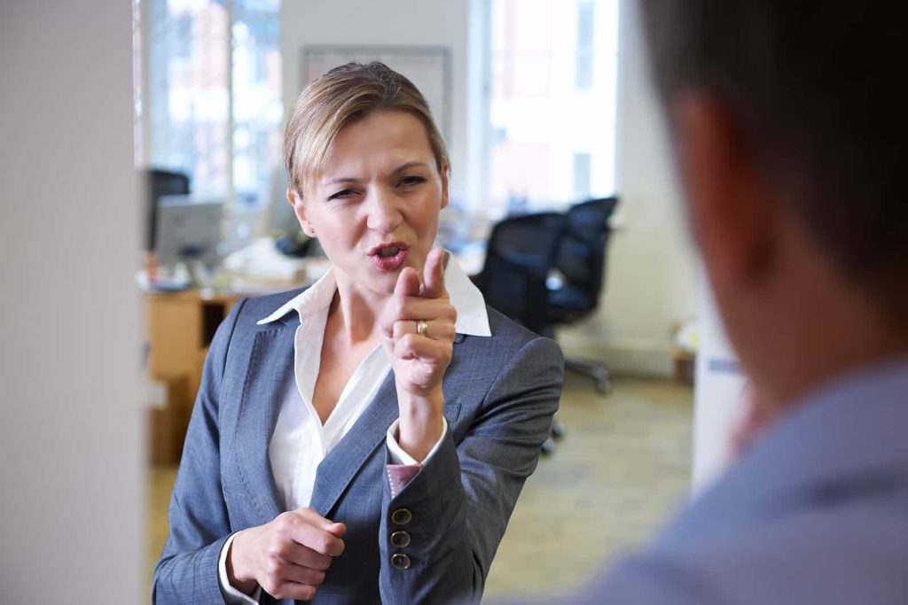 A woman in a business suit stands in an office, with a serious expression and pointing a finger at another person whose back is turned to the camera.