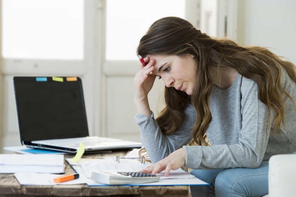 A woman sits at a cluttered table with an open laptop, papers, and a calculator, holding her forehead with one hand and appearing stressed or worried. She is focused on the calculator, possibly doing financial calculations.