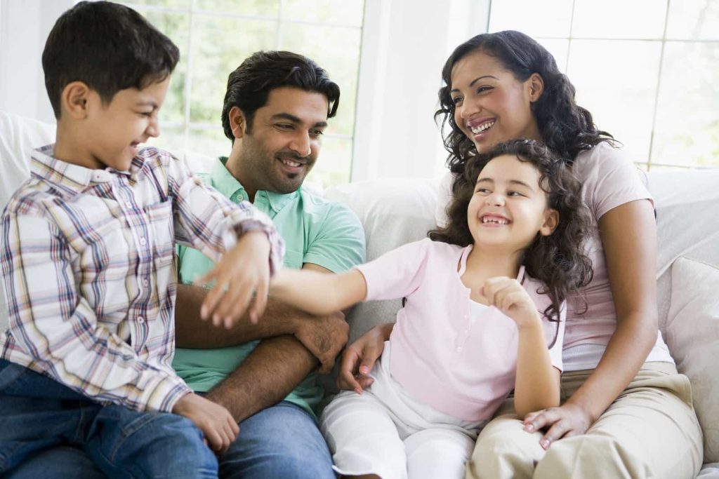 A happy family of four sits on a couch, smiling and laughing. The mother, father, daughter, and son are casually dressed. The son is playfully reaching out towards the daughter, who is giggling. A bright window in the background illuminates the scene.