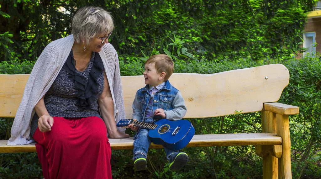 A grandmother and a young child sit on a wooden bench in a lush, green park. The child, dressed in a denim jacket and jeans, holds a small blue guitar and smiles up at the woman. The grandmother, wearing a red skirt, black top, and a gray shawl, smiles back warmly.