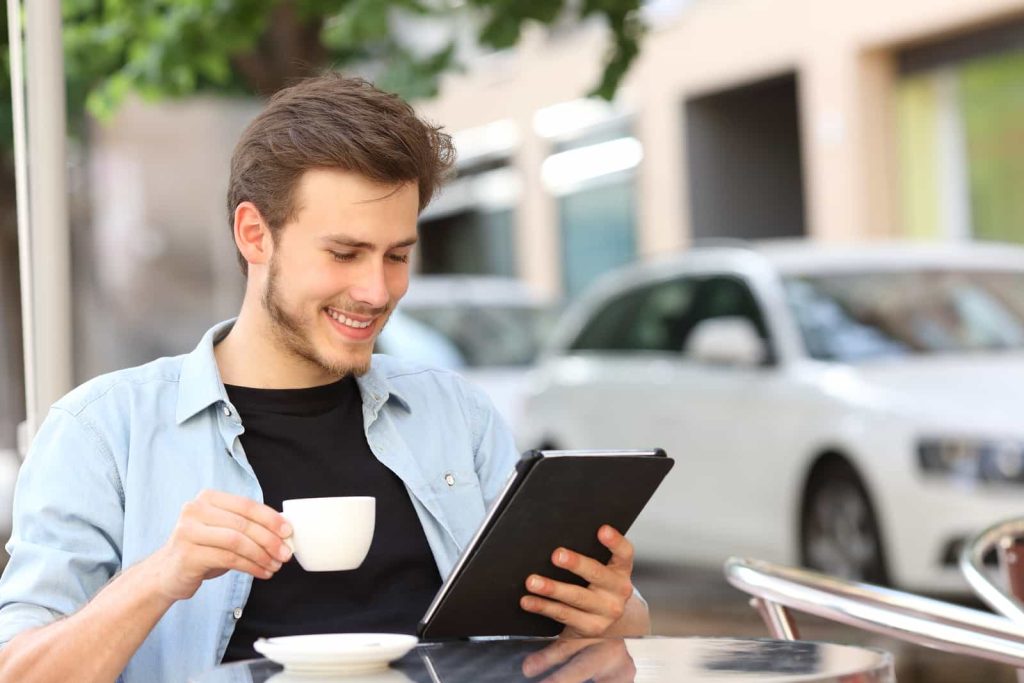 A smiling man sits at an outdoor café, holding a cup of coffee in one hand and reading on a tablet with the other. He is dressed casually in a light blue shirt. Cars and buildings are visible in the background.