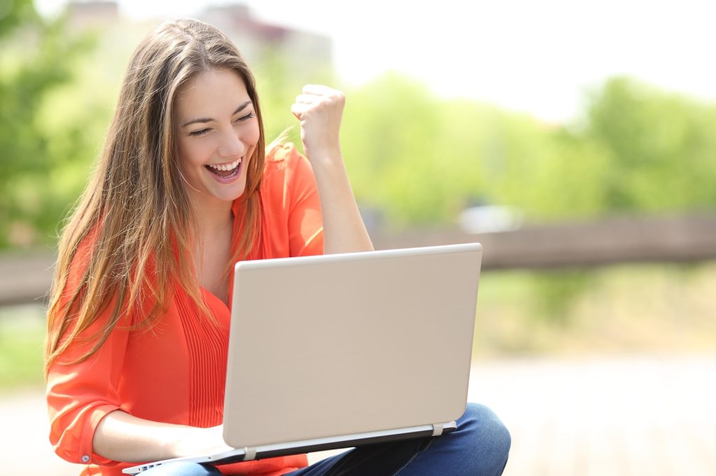 A woman with long brown hair, wearing an orange blouse, is sitting outside and looking at her laptop. She has a joyful expression and is raising her right fist in celebration.