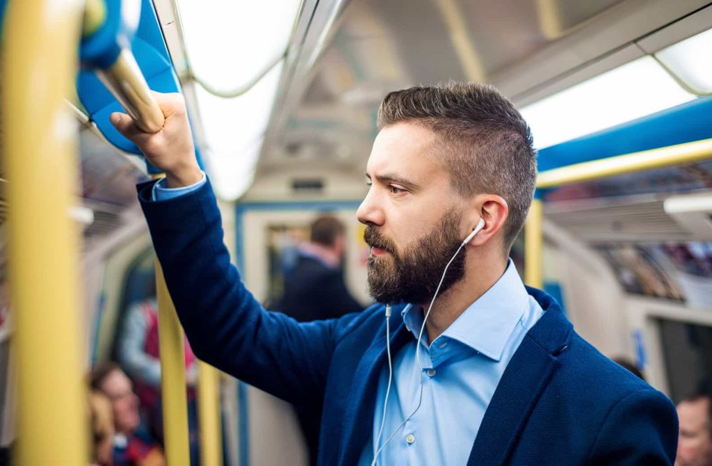 A man with a beard, wearing a blue shirt and blazer, is standing in a subway train, holding onto a rail with one hand and listening to earphones. Other passengers are seated and standing in the background. The setting appears to be an urban commuter train.